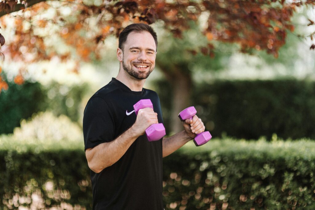 Smiling man in black shirt exercises outdoors with purple dumbbells in a park setting.