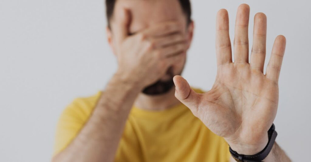 Man in yellow shirt covering face with one hand while showing a stop gesture with the other.