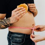 A man with tattoos holding a burger as another person measures his waistline with a tape measure.