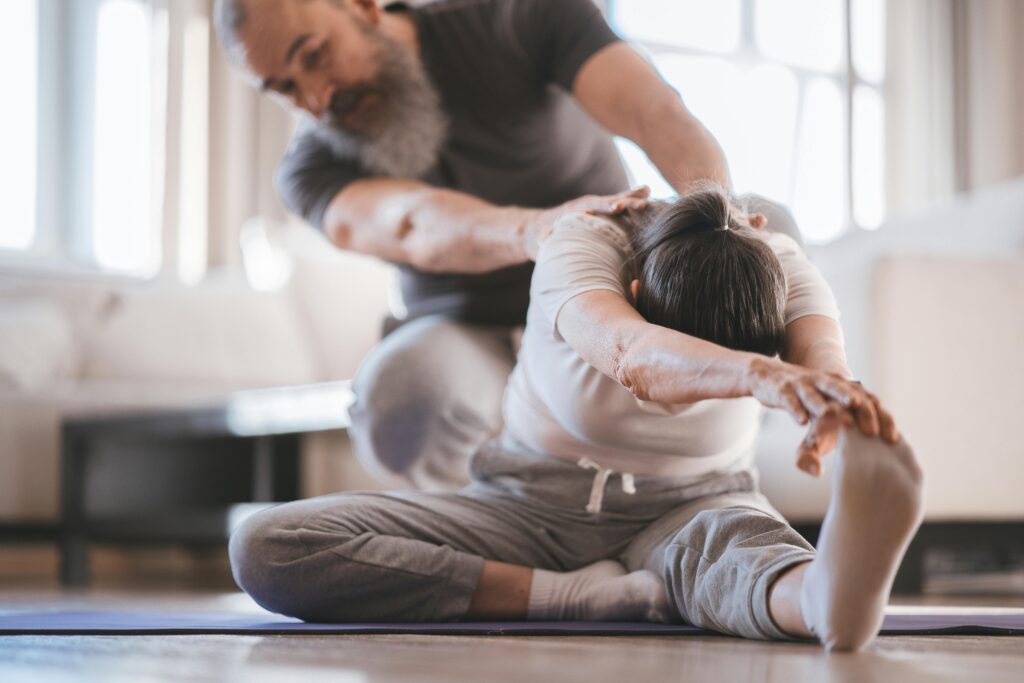 Senior couple practicing yoga at home with assistance, focusing on flexibility and wellness.