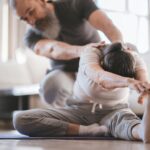 Senior couple practicing yoga at home with assistance, focusing on flexibility and wellness.