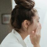 A young woman in a bathrobe follows her skincare routine, washing her face in a bathroom.