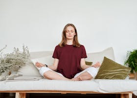 Woman meditating indoors on a sofa, exuding calmness and mindfulness, surrounded by greenery.
