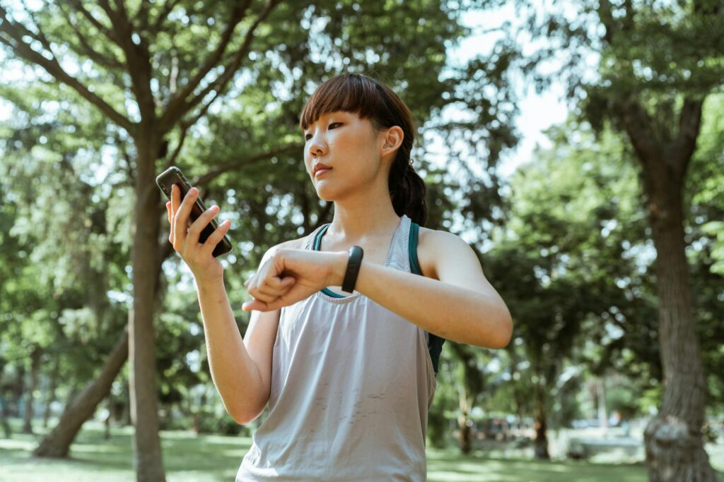 Positive concentrated young fit Asian sportswoman in activewear browsing smartphone and checking fitness bracelet while training in green sunny park