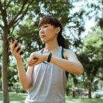 Positive concentrated young fit Asian sportswoman in activewear browsing smartphone and checking fitness bracelet while training in green sunny park
