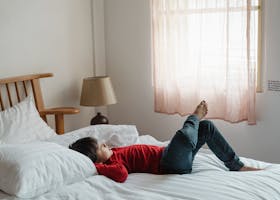 A child in a red shirt relaxes on a cozy bed in a well-lit bedroom. Perfect for lifestyle or home decor themes.