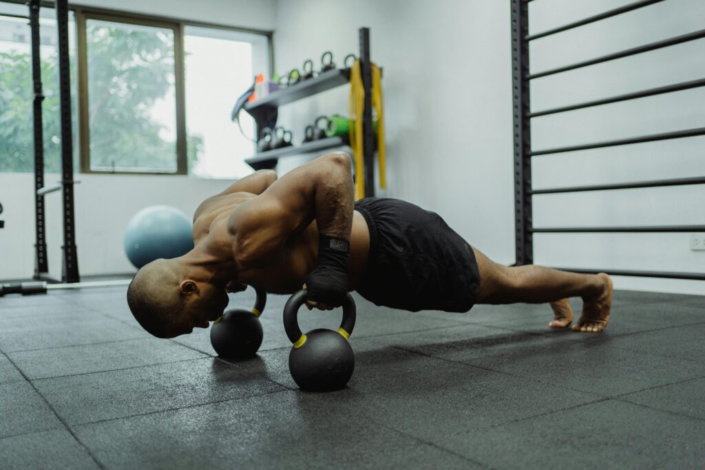 A shirtless man with a muscular build exercises in a gym, performing push-ups using kettlebells.