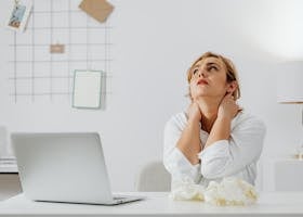 A stressed woman in white long sleeves sits at a desk with a laptop, looking upwards in an office.
