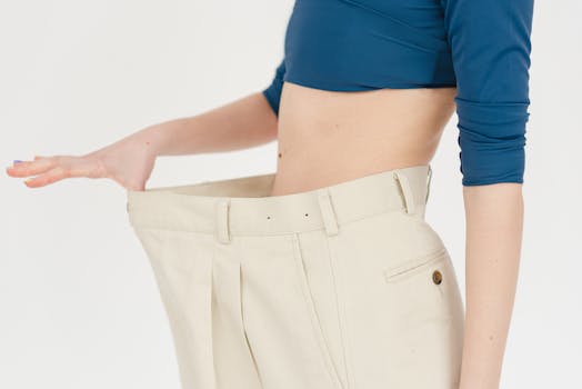 Unrecognizable slim female with bare belly wearing white oversized pants after weight loss on white background in modern light studio