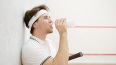 A young man resting and drinking water in a squash court, with a racket by his side.