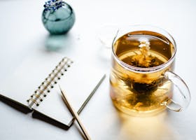 Close-up of flowering tea in a transparent pot alongside a notebook and pencil, offering a peaceful tabletop scene.