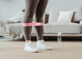 Close-up of a woman in sport leggings using a pink resistance band for leg exercises indoors.