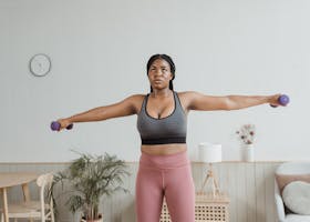 Black woman in activewear performing arm exercises indoors with dumbbells.