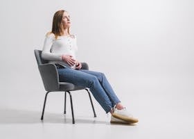 A pregnant woman in a white shirt sitting on a chair, gazing thoughtfully.