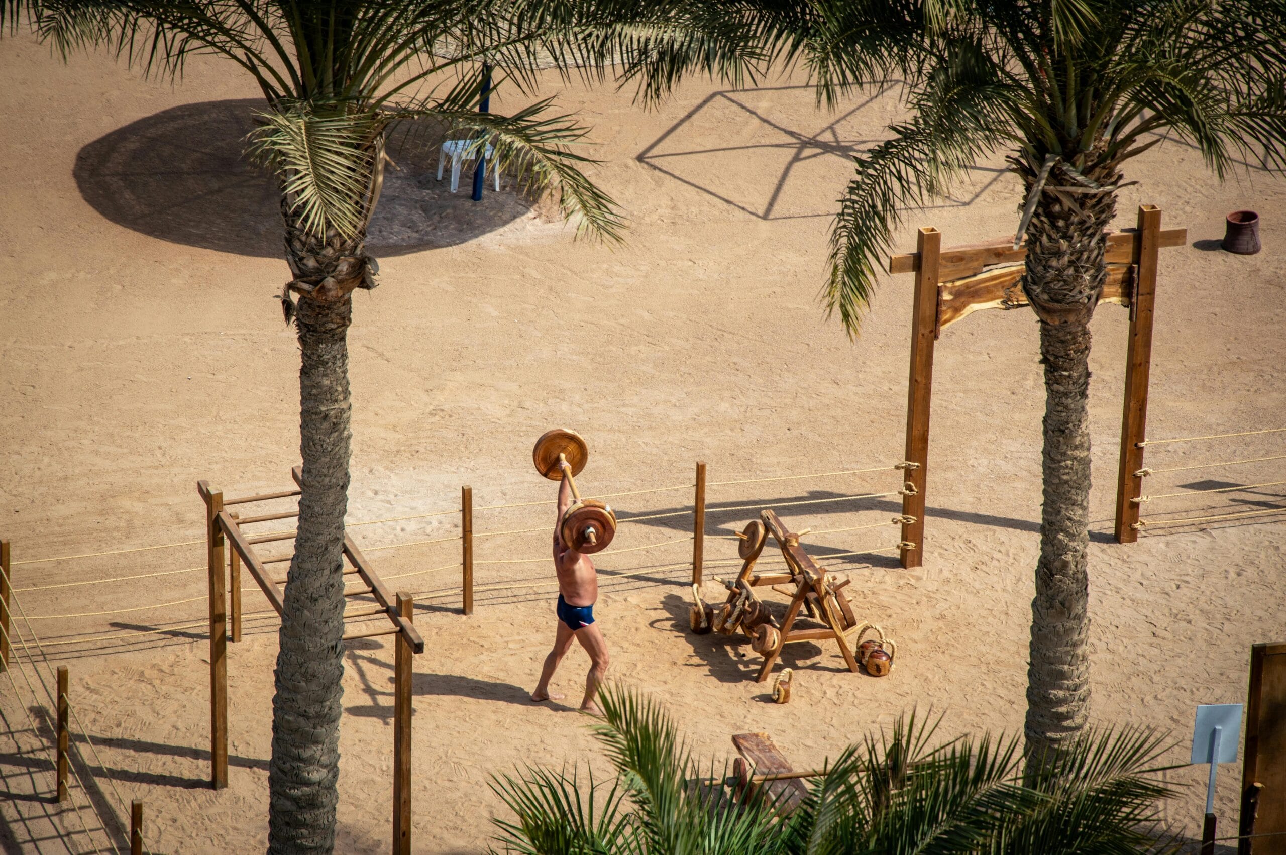 Man lifting weights in a sandy outdoor gym among palm trees, viewed from above.