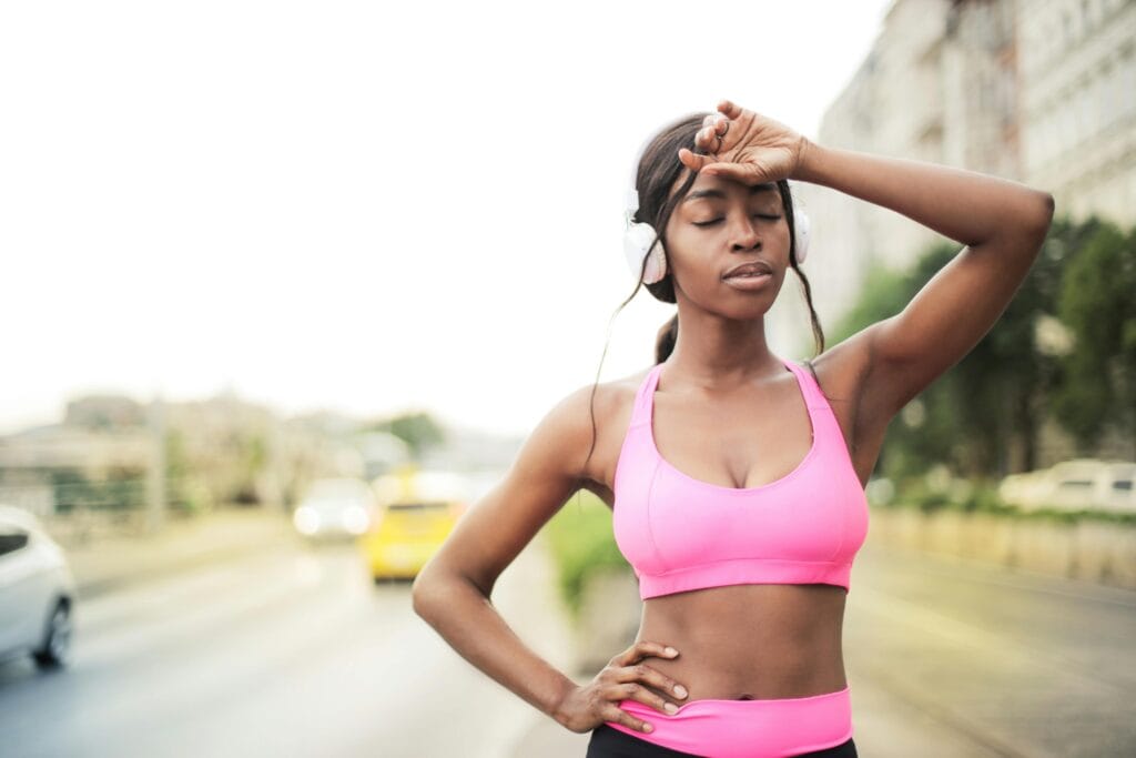 Fit black woman in sports bra resting after outdoor workout with headphones on a sunny day.