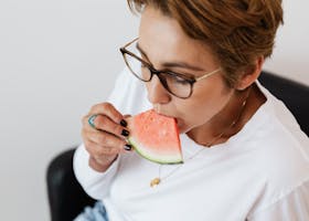 From above of crop short haired lady in casual wear and eyeglasses eating delicious juicy watermelon sitting in leather armchair