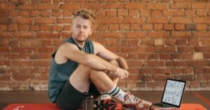 A man sits on an exercise mat with a laptop showing workout routines, suggesting a fitness regimen.