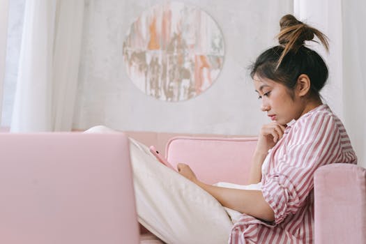 Asian woman using her phone on a pink sofa at home, relaxed and thoughtful in a cozy setting.