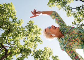 Senior woman expressing joy while dancing under green trees on a sunny summer day.