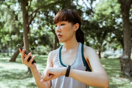 Serious young Asian female in sportswear using smartphone and smart watch in park on sunny summer day