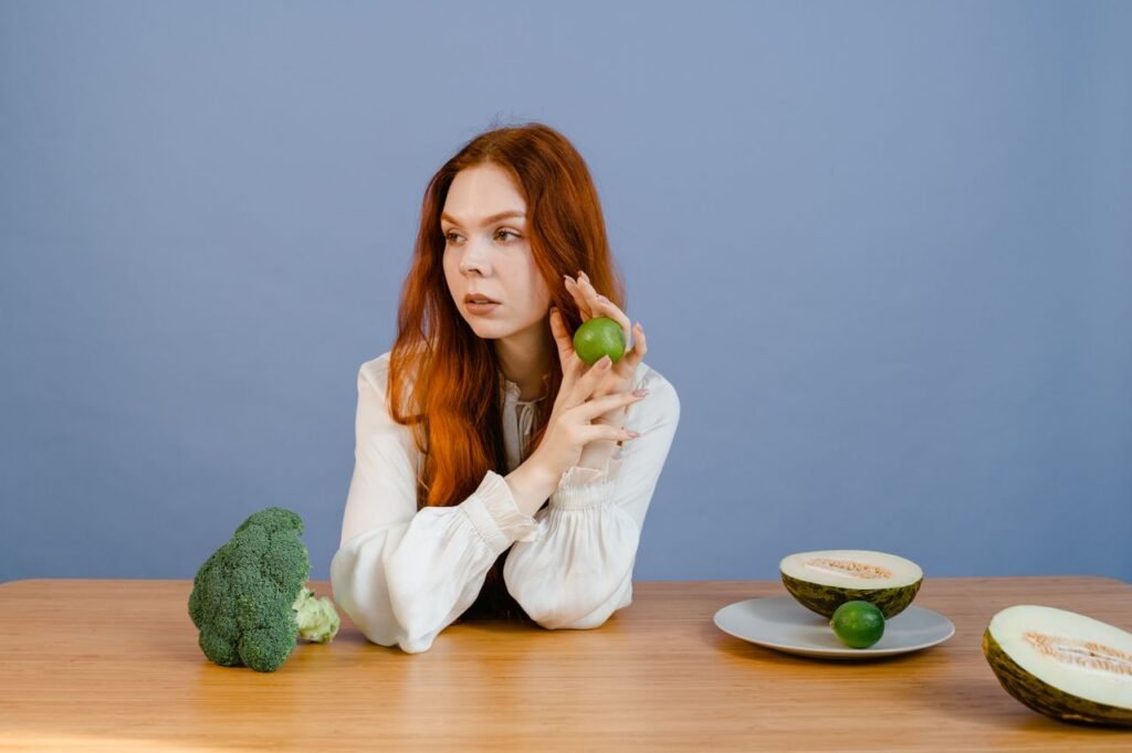 Caucasian woman sitting at table holding lime with broccoli and melon.