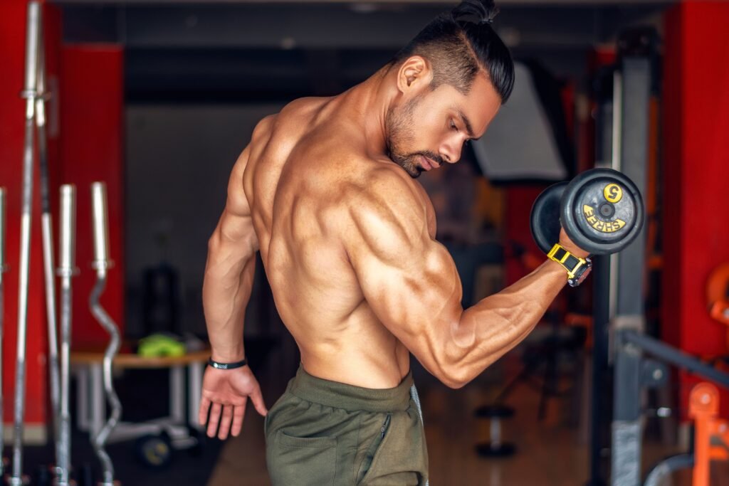 A fit, shirtless man showcasing biceps while lifting a dumbbell in a gym setting.